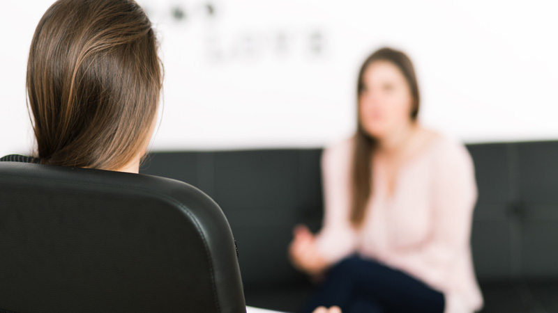 A woman sitting at home in a chair talking to another woman.